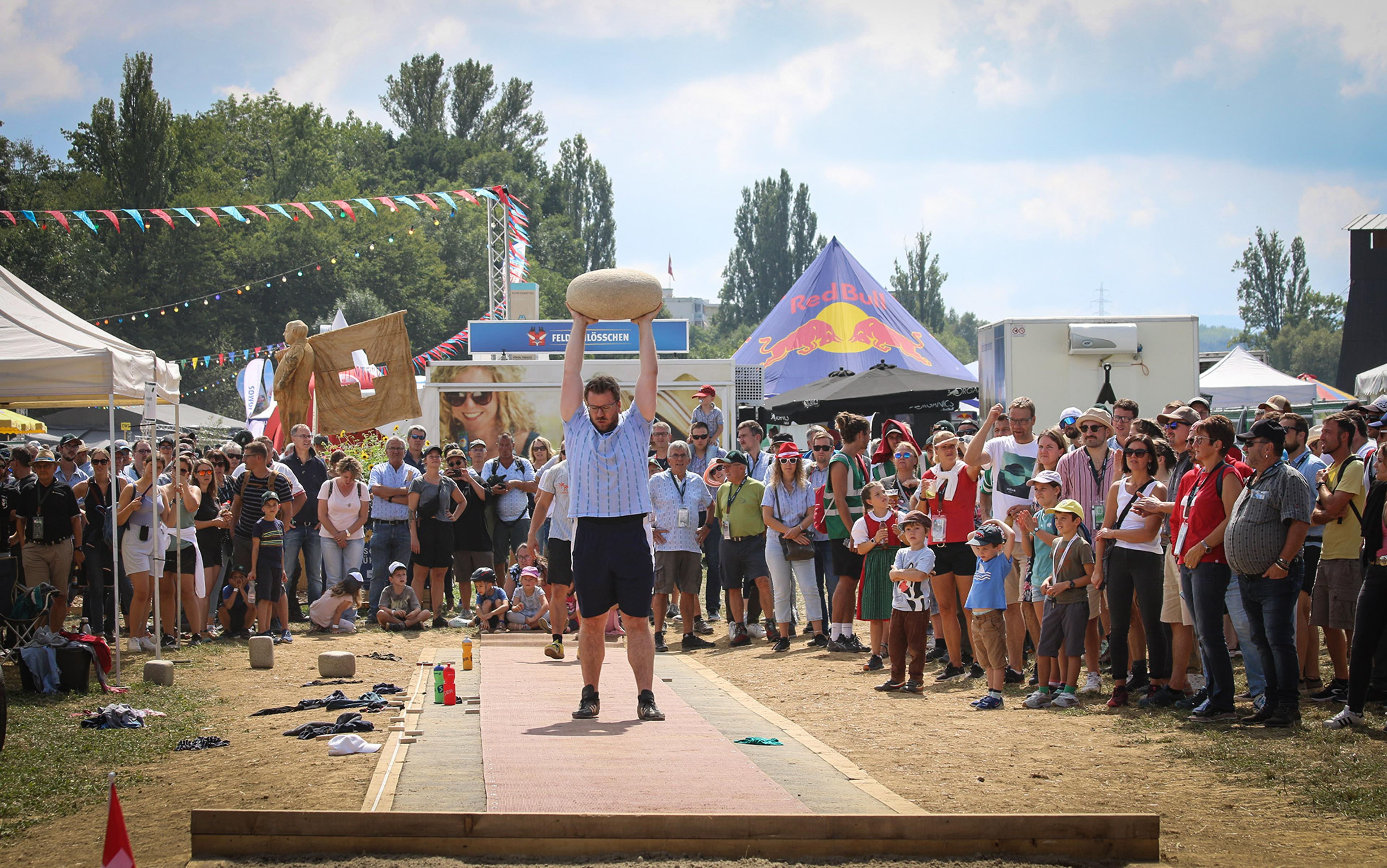 A man in tradional long shorts and striped shirt holds a huge heavy stone abvove his head as a crowd looks on