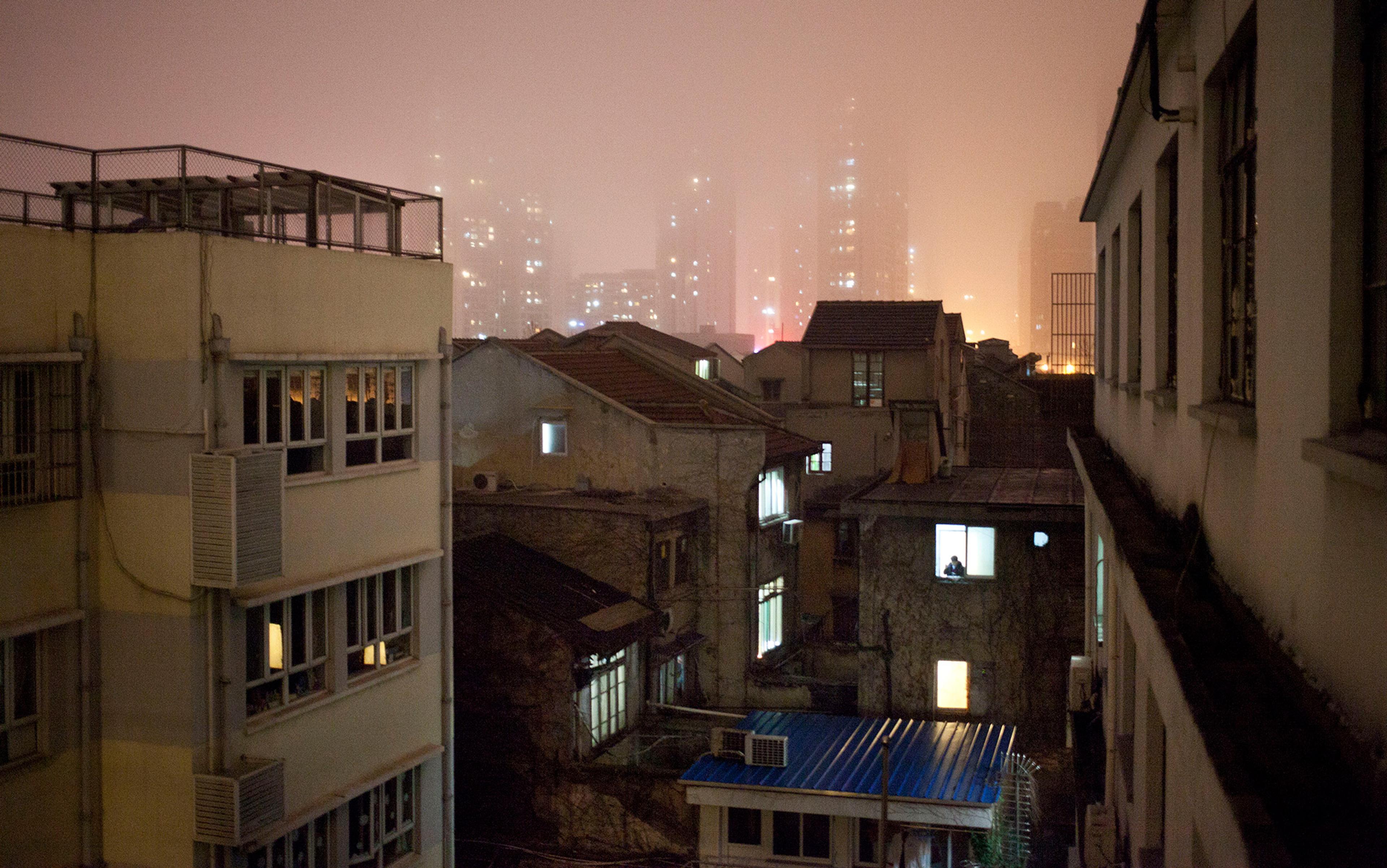 A figure is silhouetted in an older style apartment block window at night. In the distance are taller modern tower block apartments