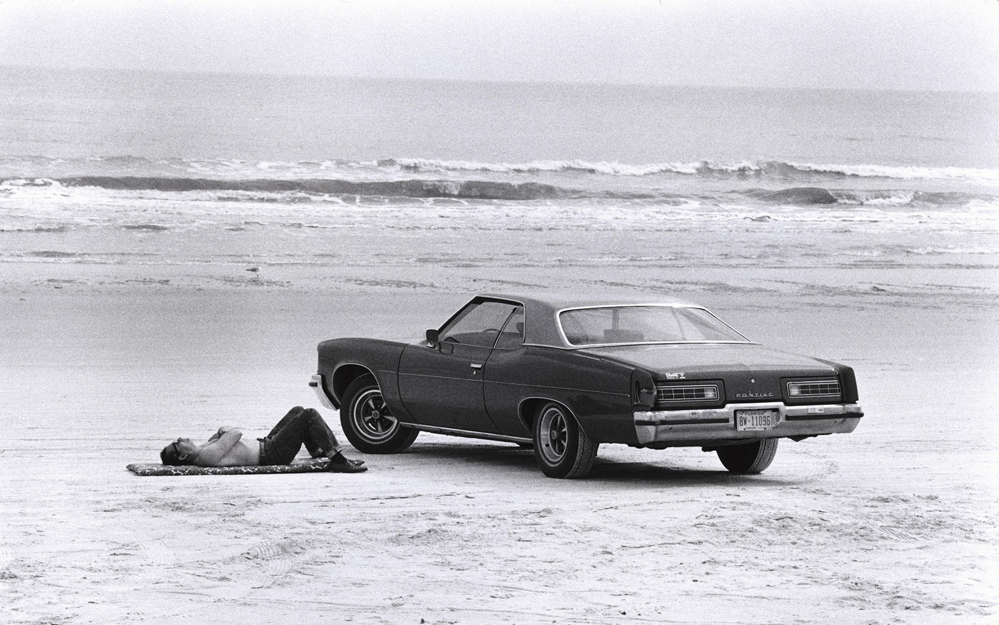 A shirtless man in jeans lies sleeping beside his 1970s era car on the sandy beach